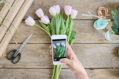 Directly above view of unrecognizable woman taking picture of pretty pink tulip bouquet on wooden table with smartphone