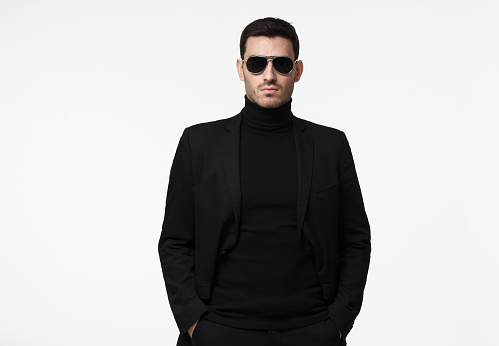 Half-length portrait of young handsome European guy isolated on white background dressed in total black with turtleneck, sunglasses and jacket, looking serious and suspicious, reserved and reticent