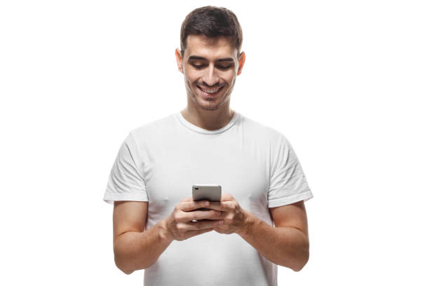 istock Young man in blank tshirt standing isolated on white background looking attentively at screen of cellphone, browsing web pages and smiling nicely while chatting with friends 1022250216