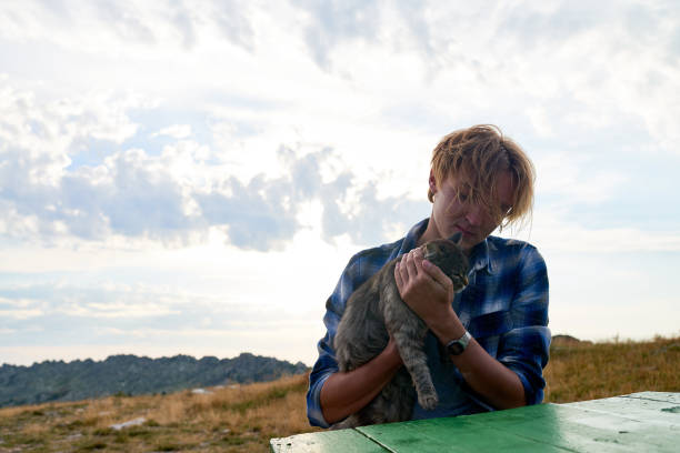 Young Caucasian male hiker caressing cute gray cat while sitting at wooden table outdoors on cloudy sky background