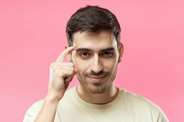 closeup portrait of young european caucasian man pictured isolated on pink background pressing finger to temple as if making viewer think more about offer or analyze information better for their good - outdoor lifestyle imagens e fotografias de stock