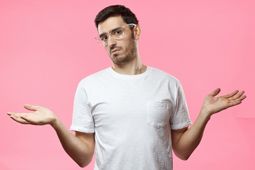 I don't know. Young man isolated on pink background being at a loss, showing helpless gesture with arm and hands, mouth curved as if he does not know what to do