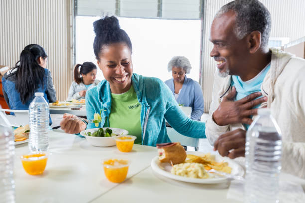 Female volunteer has lunch with man in soup kitchen Mid adult African American female volunteer has lunch with a senior African American man in a soup kitchen. The woman is laughing at a joke the man told her. community center food stock pictures, royalty-free photos & images