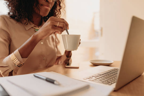 Woman holding a cup of coffee while working on laptop at home Businesswoman working on laptop computer sitting at home holding a coffee cup in hand. Woman entrepreneur stirring her coffee with spoon while working on laptop. stirring stock pictures, royalty-free photos & images