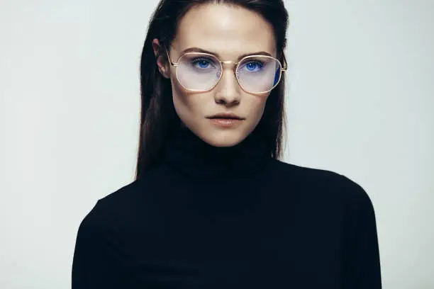 Close up portrait of woman wearing glasses staring with an intense expression. Female model in black dress on grey background.