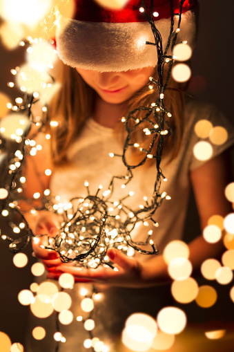 Beautiful little girl wearing Santa's hat and holding bunch of Christmas lights; child setting up Christmas decorations and waiting for Santa