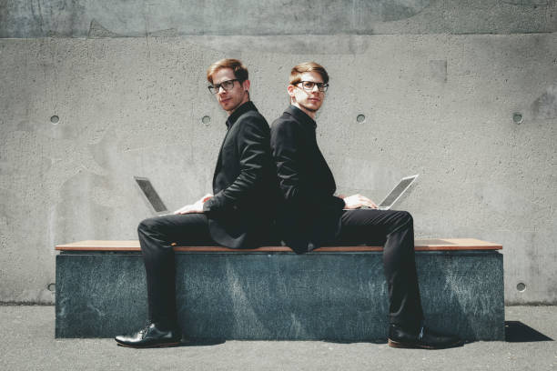 twin brothers shoulder on shoulder working twin brothers with black suits and glasses sitting in urban surrounding shoulder on shoulder with their laptops and working global wireless. two men same clothes stock pictures, royalty-free photos & images