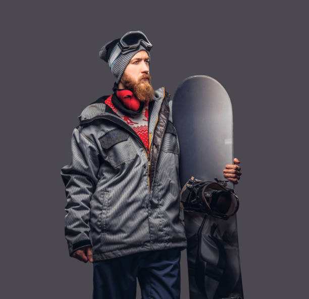 istock Brutal redhead snowboarder with a full beard in a winter hat and protective glasses dressed in a snowboarding coat posing with snowboard at a studio, looking away. Isolated on the gray background. 1022405024