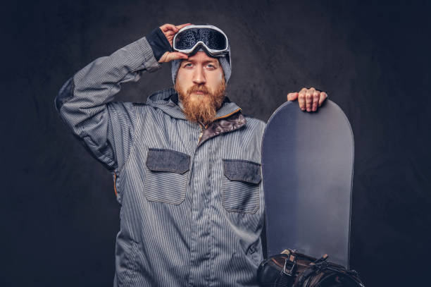 istock Brutal redhead snowboarder with a full beard in a winter hat and protective glasses dressed in a snowboarding coat posing with snowboard at a studio. Isolated on gray background. 1022407464