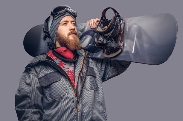 istock Brutal redhead snowboarder with a full beard in a winter hat and protective glasses dressed in a snowboarding coat posing with snowboard at a studio, looking away. Isolated on the gray background. 1022407672