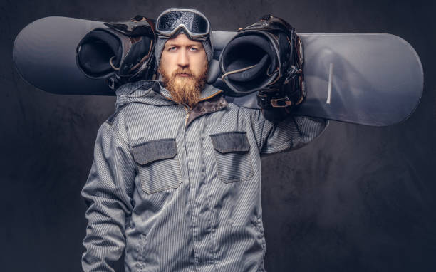 istock Brutal redhead snowboarder with a full beard in a winter hat and protective glasses dressed in a snowboarding coat posing with snowboard at a studio. Isolated on gray background. 1022408398