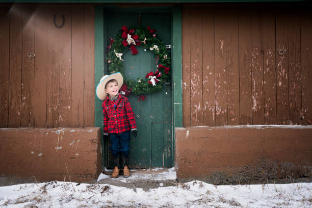 Laughing little cowboy and Christmas wreath Caucasian 4 year old boy in red plaid jacket, cowboy hat, jeans, cowboy boots and gloves is laughing in front of a Christmas wreath hanging on a green door of an old barn on his ranch during winter, Livingston, Montana, USA checkered cowboy boots stock pictures, royalty-free photos & images
