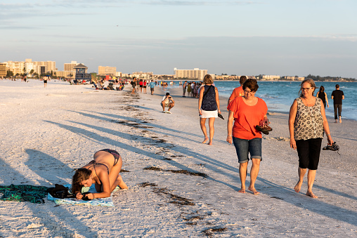 Sarasota, USA - April 27, 2018: Orange sunset in Siesta Key, Florida with coastline coast ocean gulf mexico on beach shore, many people walking, relaxing on towels