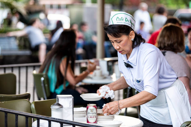 Female waitress, woman cleaning up with people sitting at table at famous, iconic Cafe Du Monde shop, serving beignet powdered sugar donuts, beer New Orleans, USA - April 23, 2018: Female waitress, woman cleaning up with people sitting at table at famous, iconic Cafe Du Monde shop, serving beignet powdered sugar donuts, beer beignet stock pictures, royalty-free photos & images