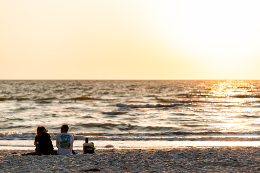 Naples, USA - April 29, 2018: Florida vintage orange golden yellow soft sunset sunlight in gulf of Mexico with two people young couple sitting on sand drinking tea, romantic