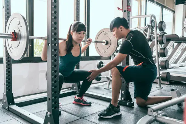Woman doing squats with barbell. The personal trainer helping her flexing muscles in Smith machine in gym.