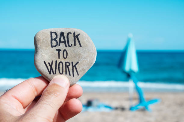 folded beach umbrella and text back to work closeup of the hand of a young caucasian man on the beach, in front of a folded beach umbrella and the ocean in the background, holding a stone with the text back to work written in it endland stock pictures, royalty-free photos & images