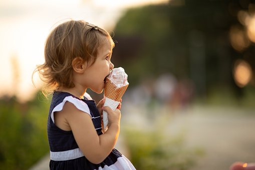 Happy preschool girl eating colorful ice cream in waffle cone on sunny summer day. Little toddler child eat icecream dessert. Sweet food on hot warm summertime days. Bright light, colorful ice-cream.