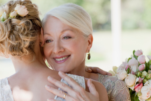 Beautiful blonde woman in white dress on her wedding day before getting married, her mother does her makeup.