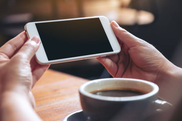 istock Mockup image of hands holding and using a white mobile phone with blank black screen horizontally for watching with coffee cup on wooden table 1022842236