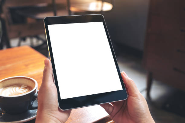 istock Mockup image of hands holding black tablet pc with blank white screen with coffee cup on wooden table in cafe 1022842286