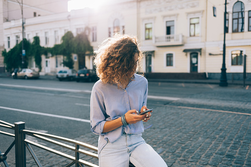 Unrecognizable curly young woman texting on smart phone sitting on parapet near european paving stone road wearing blue shirt and jeans on summer day, sun flare from behind.