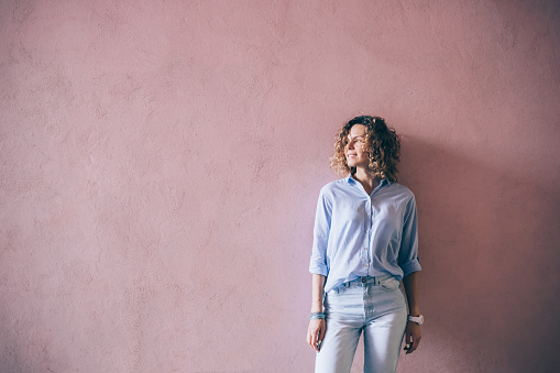 Happy young woman wearing blue casual outfit standing against pink wall looking aside and smile, copy space.