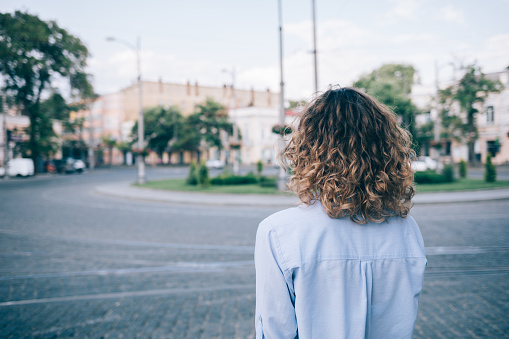 Young woman with thick curly hair looking at the european paving stone ring road. Rear view of unrecognizable female wearing blue shirt standing outside on summer day on the background of old city.