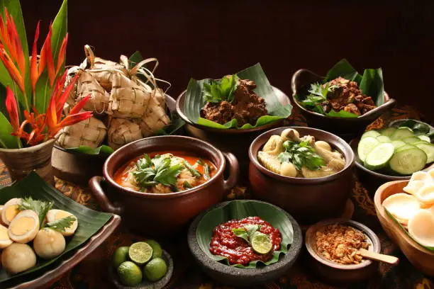 Ketupat Lebaran, the traditional celebratory dish of rice cake with rich side dishes, commonly serving during Eid celebrations. The common side dishes are Beef Rendang (Beef Dry Stew), Beef Liver in Spicy Gravy, Spicy Vegetable Soup, Chicken Curry Soup, and Marbled Soy Egg; while the condiments are fried shallots, shrimp crackers, and red chili sauce. Each dish is plated individually and arranged buffet style on a table; ready to be plated for serving. A vase of decorative banana flowers is used to decorate the table.