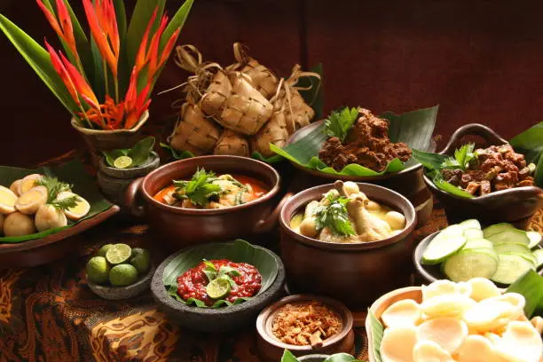 Ketupat Lebaran, the traditional celebratory dish of rice cake with rich side dishes, commonly serving during Eid celebrations. The common side dishes are Beef Rendang (Beef Dry Stew), Beef Liver in Spicy Gravy, Spicy Vegetable Soup, Chicken Curry Soup, and Marbled Soy Egg; while the condiments are fried shallots, shrimp crackers, and red chili sauce. Each dish is plated individually and arranged buffet style on a table; ready to be plated for serving. A vase of decorative banana flowers is used to decorate the table.
