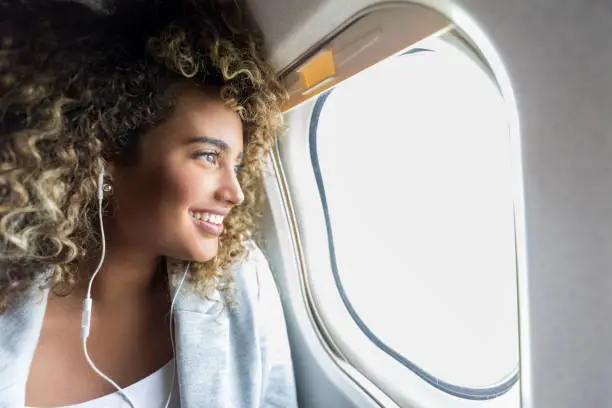Confident young mixed race woman smiles while looking through window on aircraft. She is wearing earbuds.
