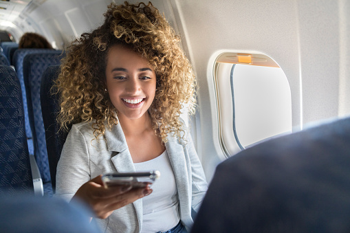 Young woman smiles while using smartphone on airplane. She is sitting in a window seat.