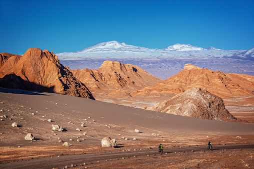 Travelers with bicycles on a track in Moon Valley, Atacama desert, snowy Andes mountain range in the background