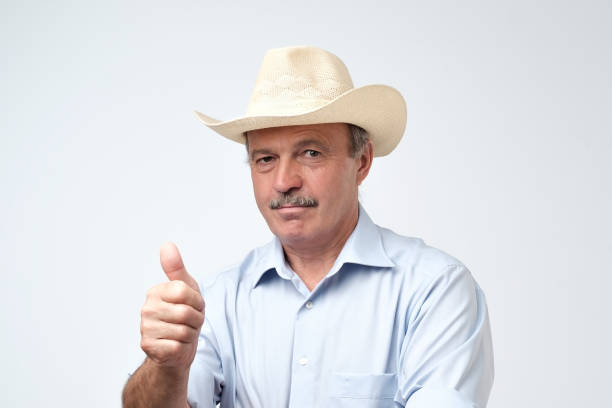 Handsome mature man in cowboy hat smiling on camera with thumb up approving your choice. Studio shoot on gray background