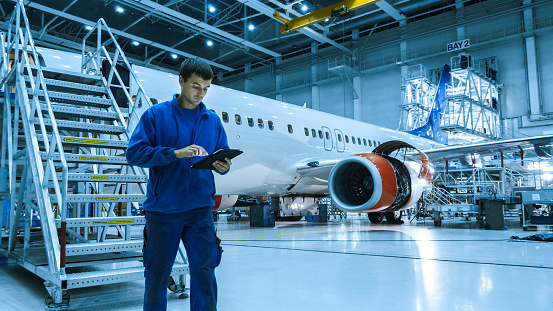 Aircraft maintenance mechanic in blue uniform is going down the stairs while using tablet in a hangar.