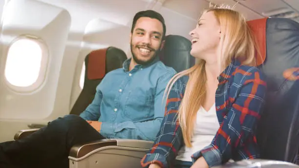 Photo of On a Commercial Plane Flight Handsome Hispanic Man Tells Funny Story to His Beautiful Blonde Girlfriend. Both Laugh. They Travel in New Airplane, with Sun Shining Through the Window.