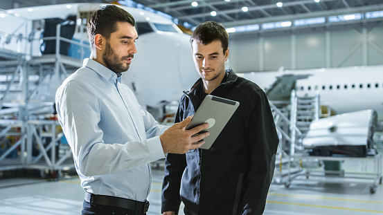 Aircraft Maintenance Worker and Engineer having Conversation. Holding Tablet.
