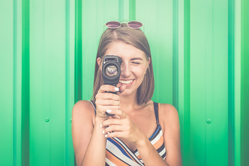 Portrait of a young smiling woman filming with retro camera isolated on green background.