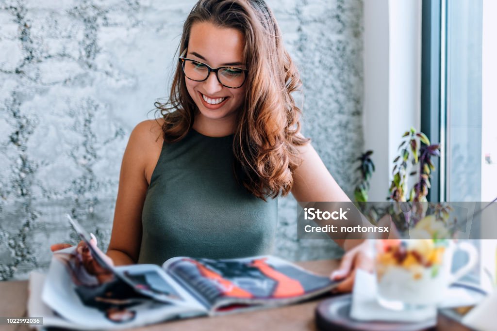 Young girl with glasses reading magazine close to window. Magazine - Publication Stock Photo