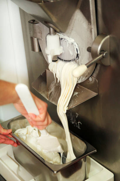 istock Female confectioner is working on ice cream maker machine. Producing vanilla and white chocolate ice cream and it falls into steel container. Industrial preparation of italian creamy ice cream. 1023031588