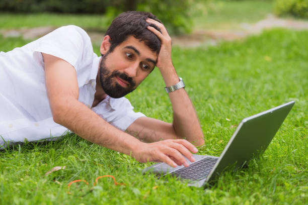 istock Young man working on his laptop while lying on the grass of the park. bored expression. 1023038668