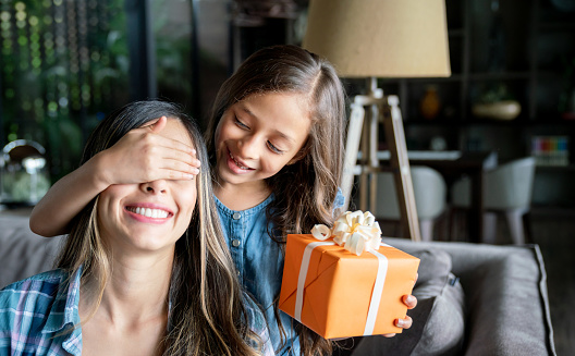 Cute little girl surprising her mom with a gift box for mother's day while covering her eyes both looking very excited