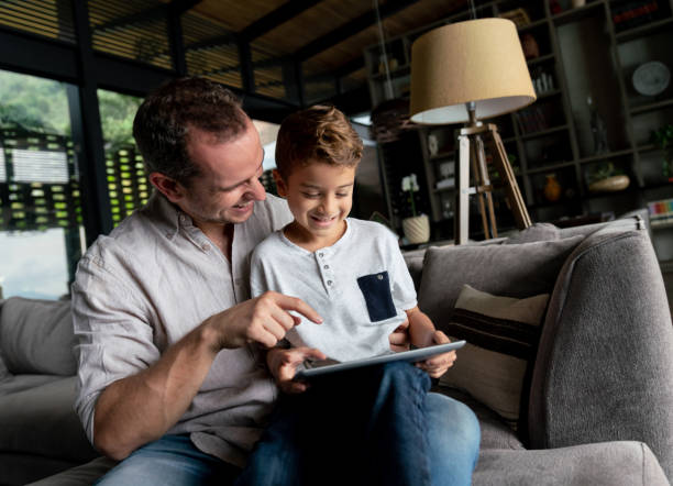istock Cute little boy sitting on his father's lap while both are playing games on tablet looking very happy 1023054218