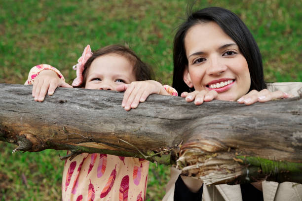 Family playing in the park Latina mom and daughter, laughing while sitting on the grass, playing hide and seek, behind a branch of a tree niñas stock pictures, royalty-free photos & images