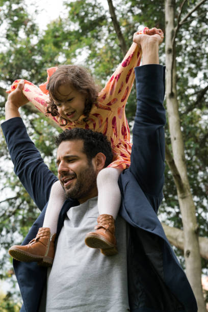 Family playing in the park Papa between 25 and 30 years old and daughter between 3 and 5 years old, playing in the park, being very happy, while papa holds her little girl on the shoulders and both have open arms niñas stock pictures, royalty-free photos & images