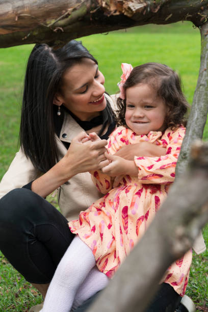 Family playing in the park Latina mom and daughter, laughing while sitting on the grass, playing tickling, in front of a tree branch niñas stock pictures, royalty-free photos & images