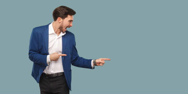 handsome amazed bearded man in blue suit standing and pointing at background and screaming and looking at copy space. - business men humor macho imagens e fotografias de stock