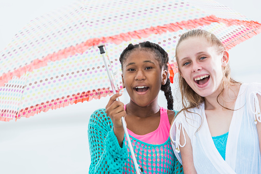 Two multi-ethnic girls on the beach standing under a shady umbrella, laughing and looking at the camera. The African-American girl is 13 years old and her friend is 12.