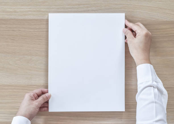 istock Blank A4 size book cover mockup template top view in businessman's hand with page front side on white surface flat lay on wood table 1023182210