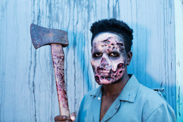 A young African-American man in his 20s dressed up as a zombie in a halloween haunted house, holding an axe, staring at the camera.